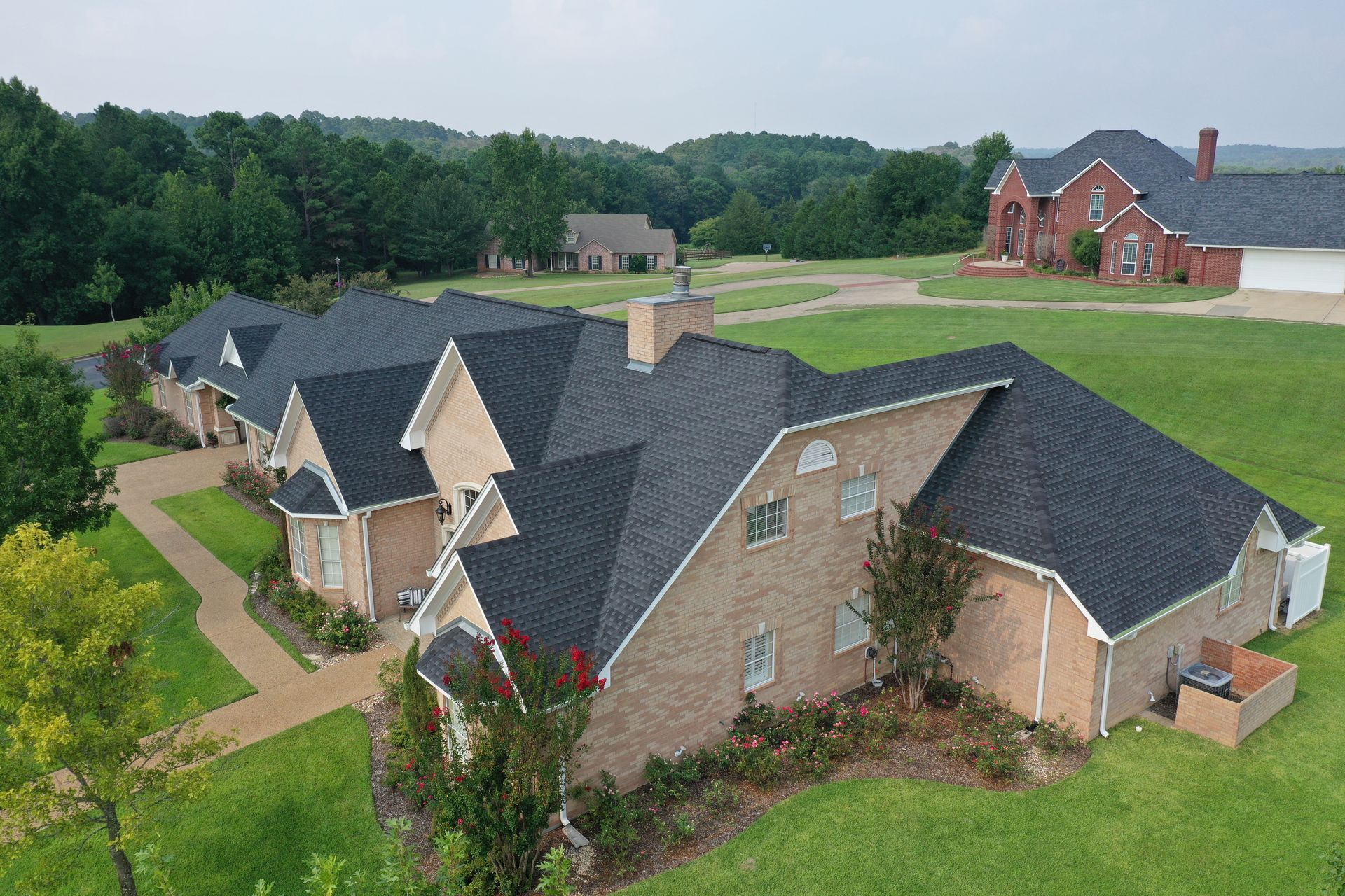 Aerial view of a large brick suburban house with dark roofs, green lawn, and trees nearby.