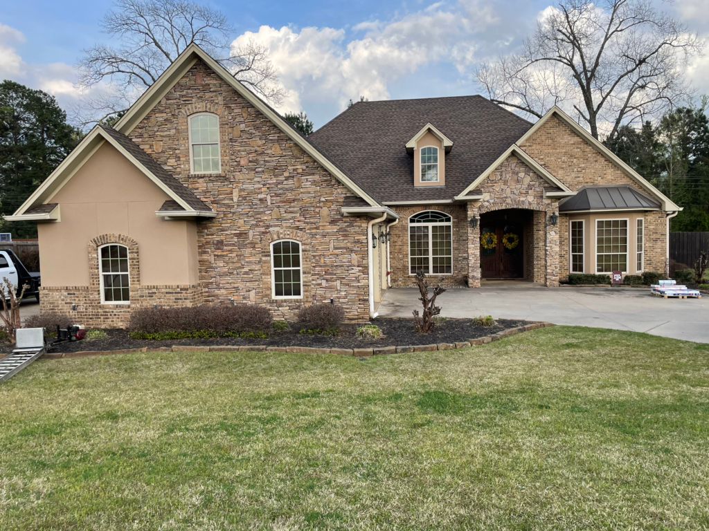 Large brick suburban house with a front porch, driveway, and landscaped lawn under a cloudy sky