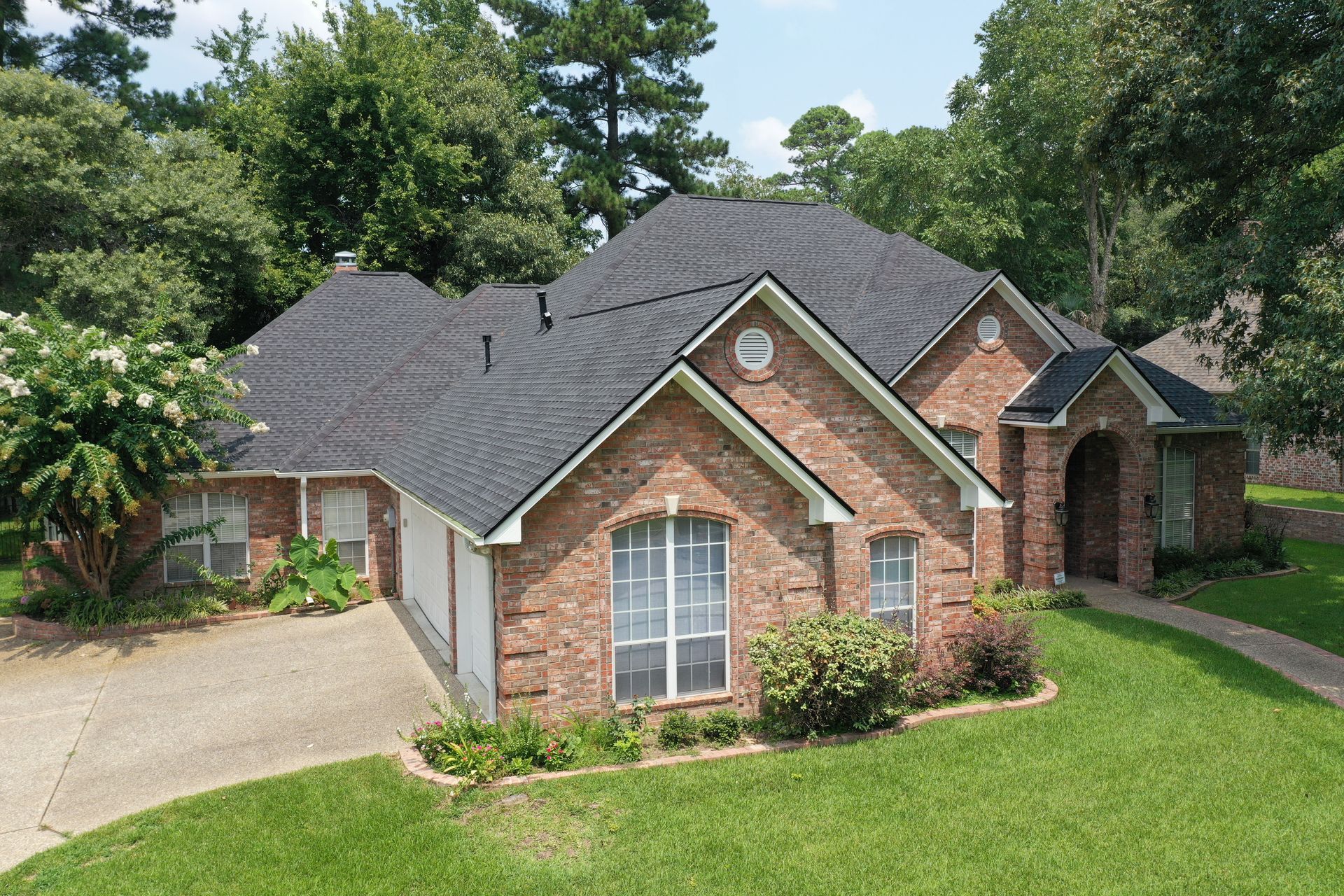 Brick suburban house with gray roof, white-trim windows, driveway, and green lawn surrounded by trees