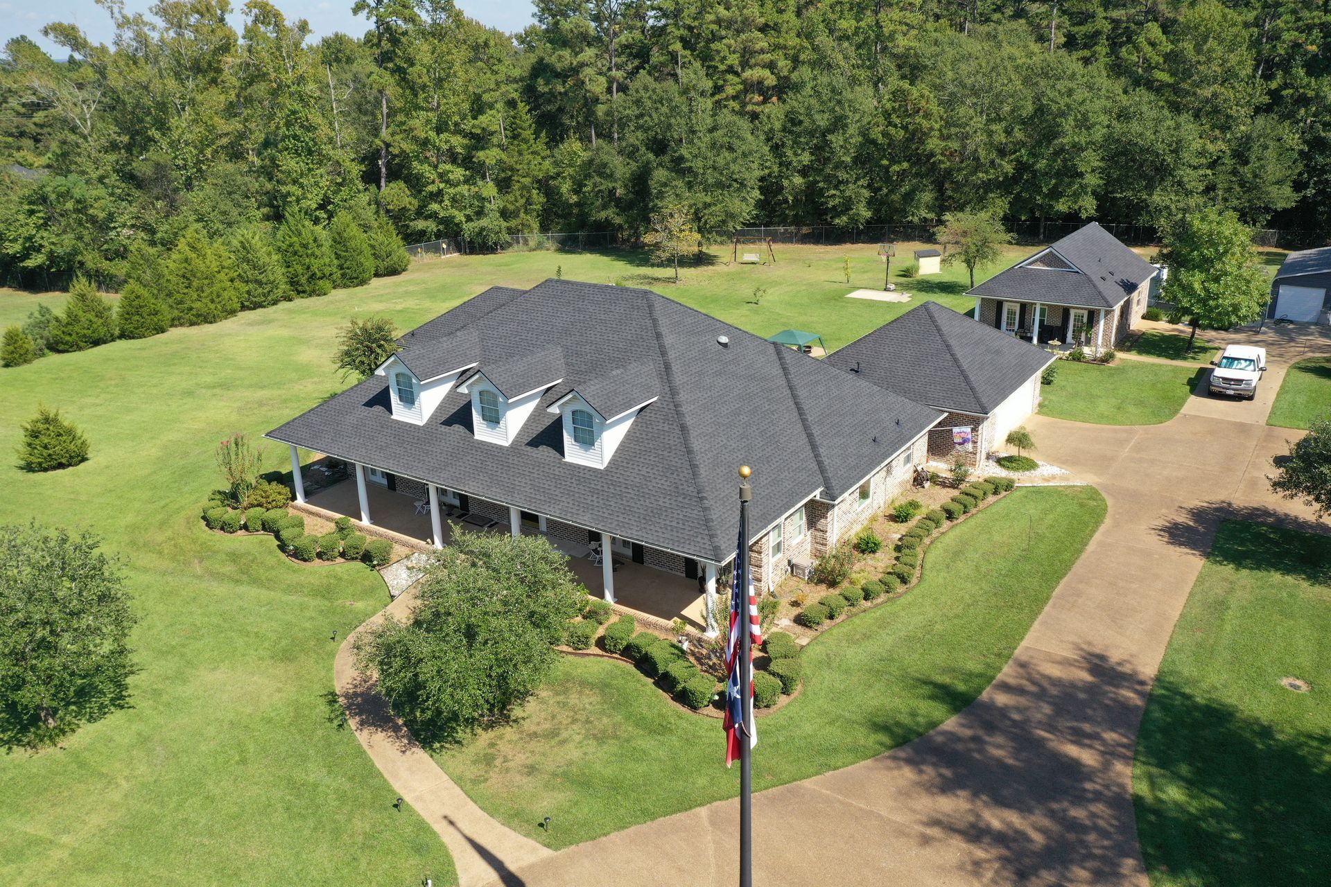 Aerial view of a house with a dark roof, surrounded by green lawn and trees, with a driveway and flagpole.