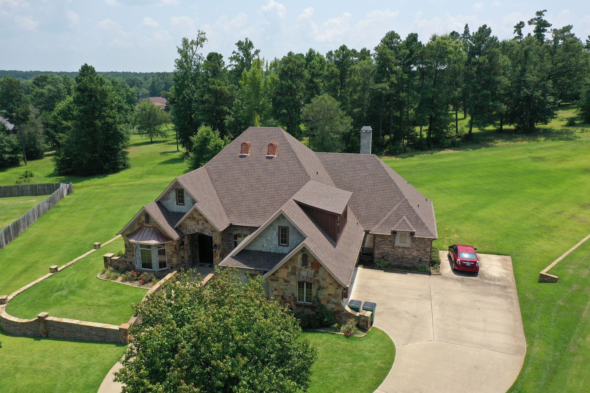 Large stone house with a brown roof and driveway, surrounded by a grassy yard and trees.