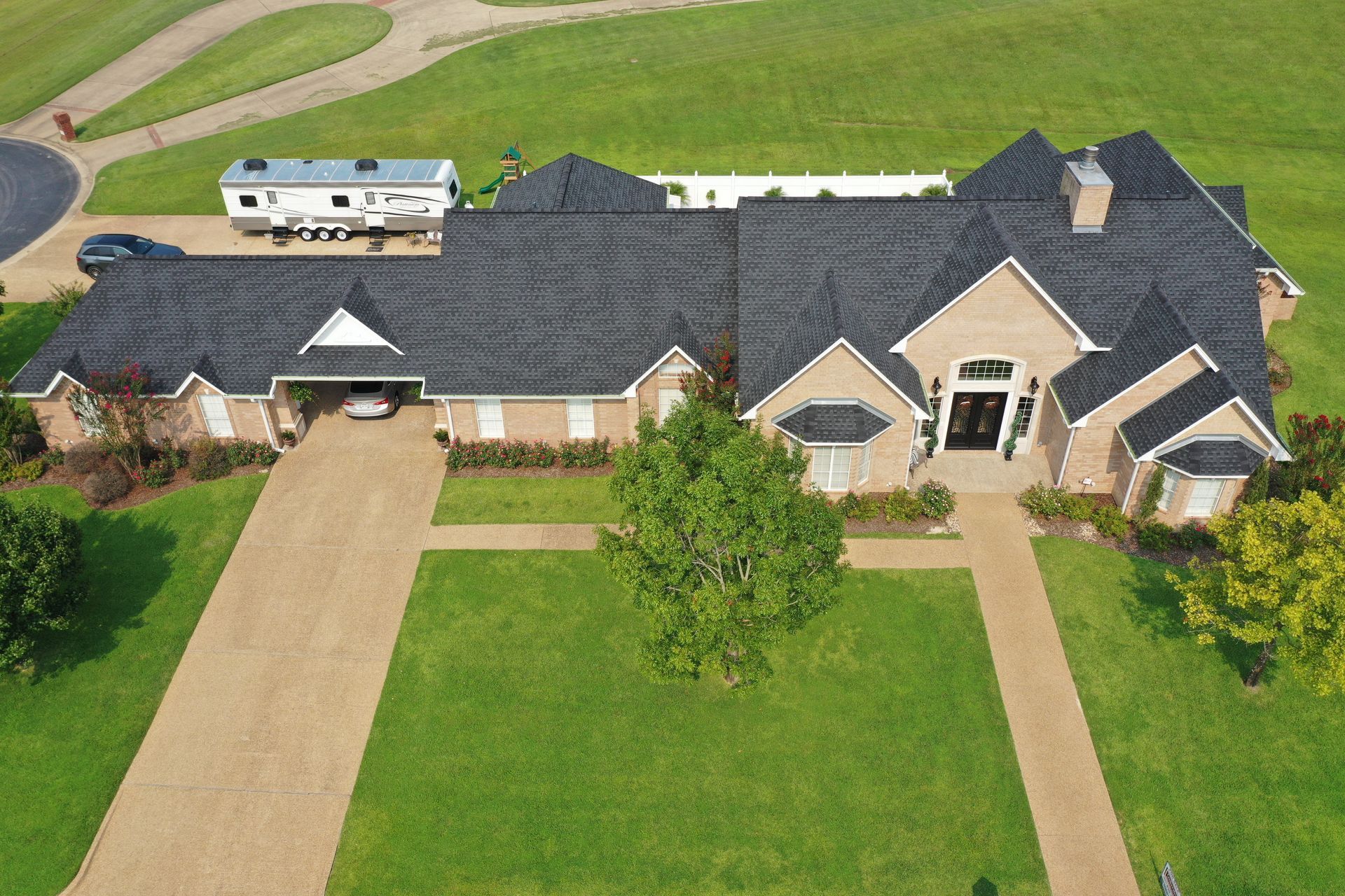 Aerial view of a large beige house with a dark roof, two driveways, and a green lawn.