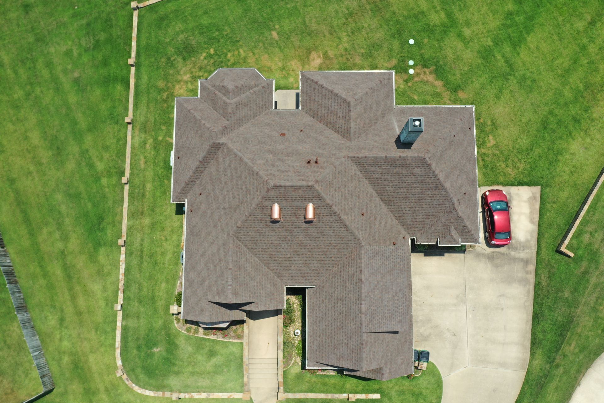 Aerial view of a house with a gray roof, driveway, and a red car beside it, surrounded by green lawn
