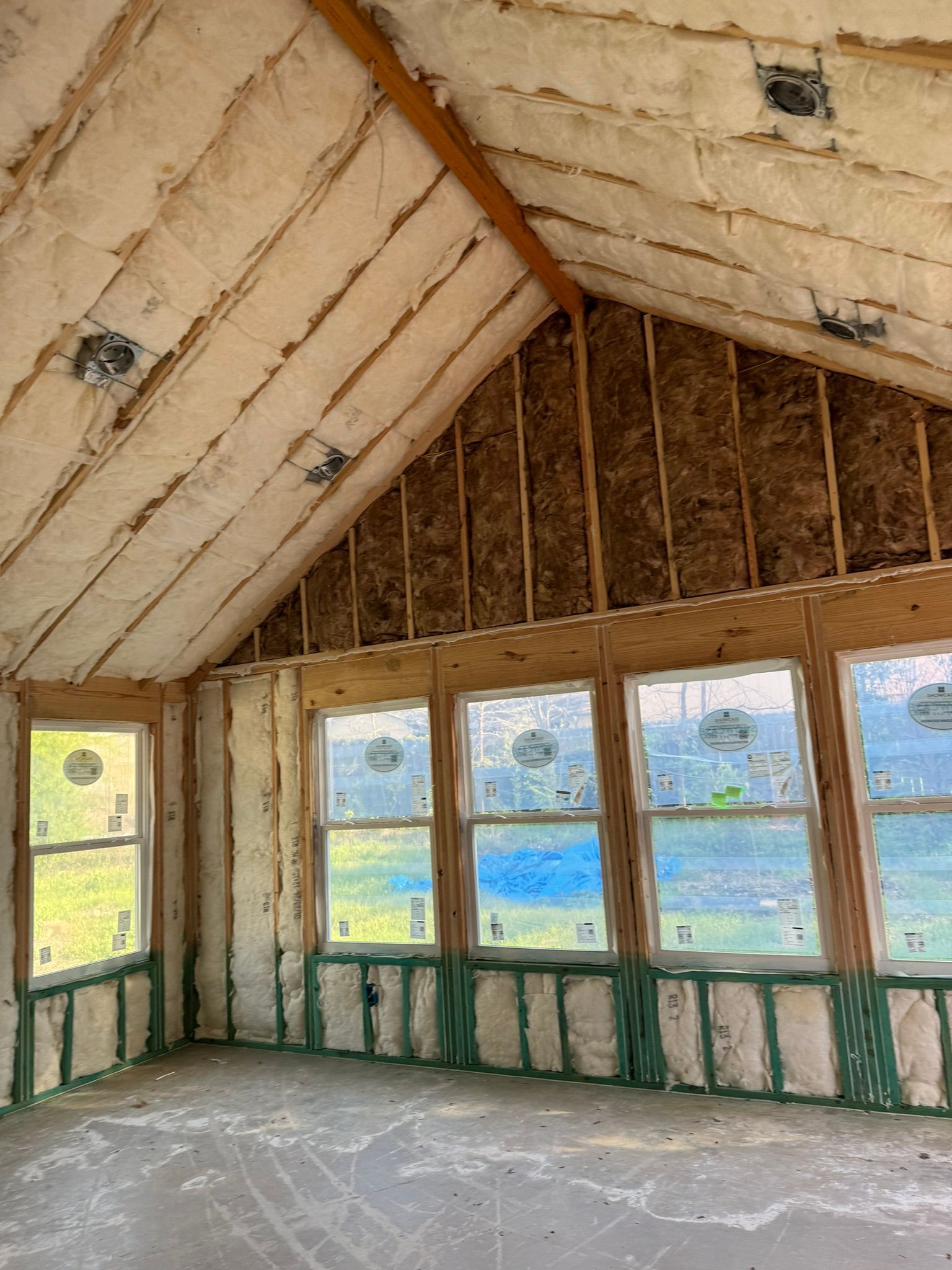 Interior of an unfinished room with exposed insulation, sloped ceiling, and windows overlooking greenery