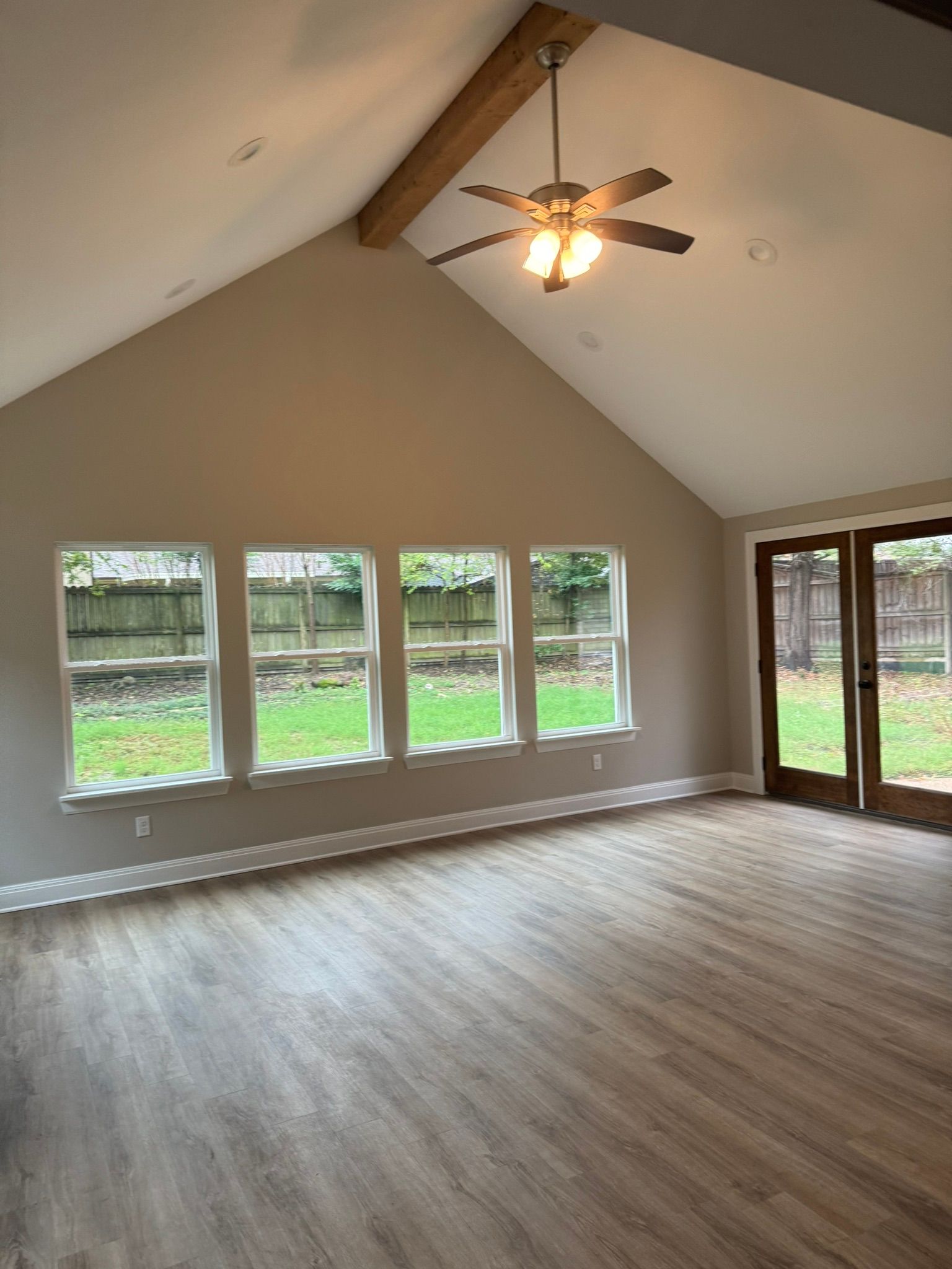 Empty living room with vaulted ceiling, ceiling fan, wide windows, and sliding glass doors to a backyard.