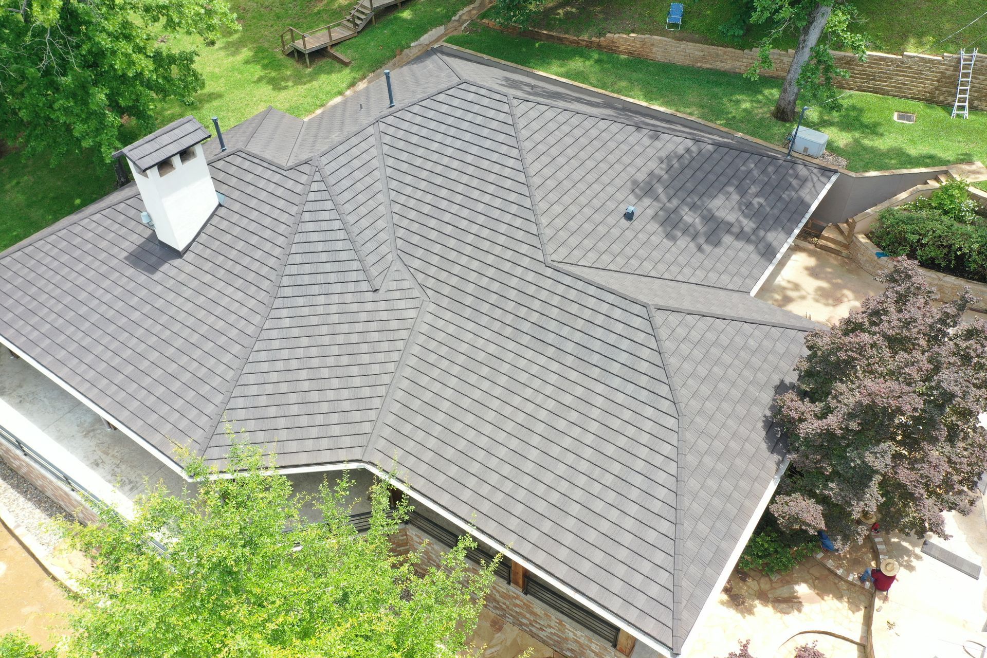 Aerial view of a house with a gray shingle roof and white chimney, surrounded by green trees.