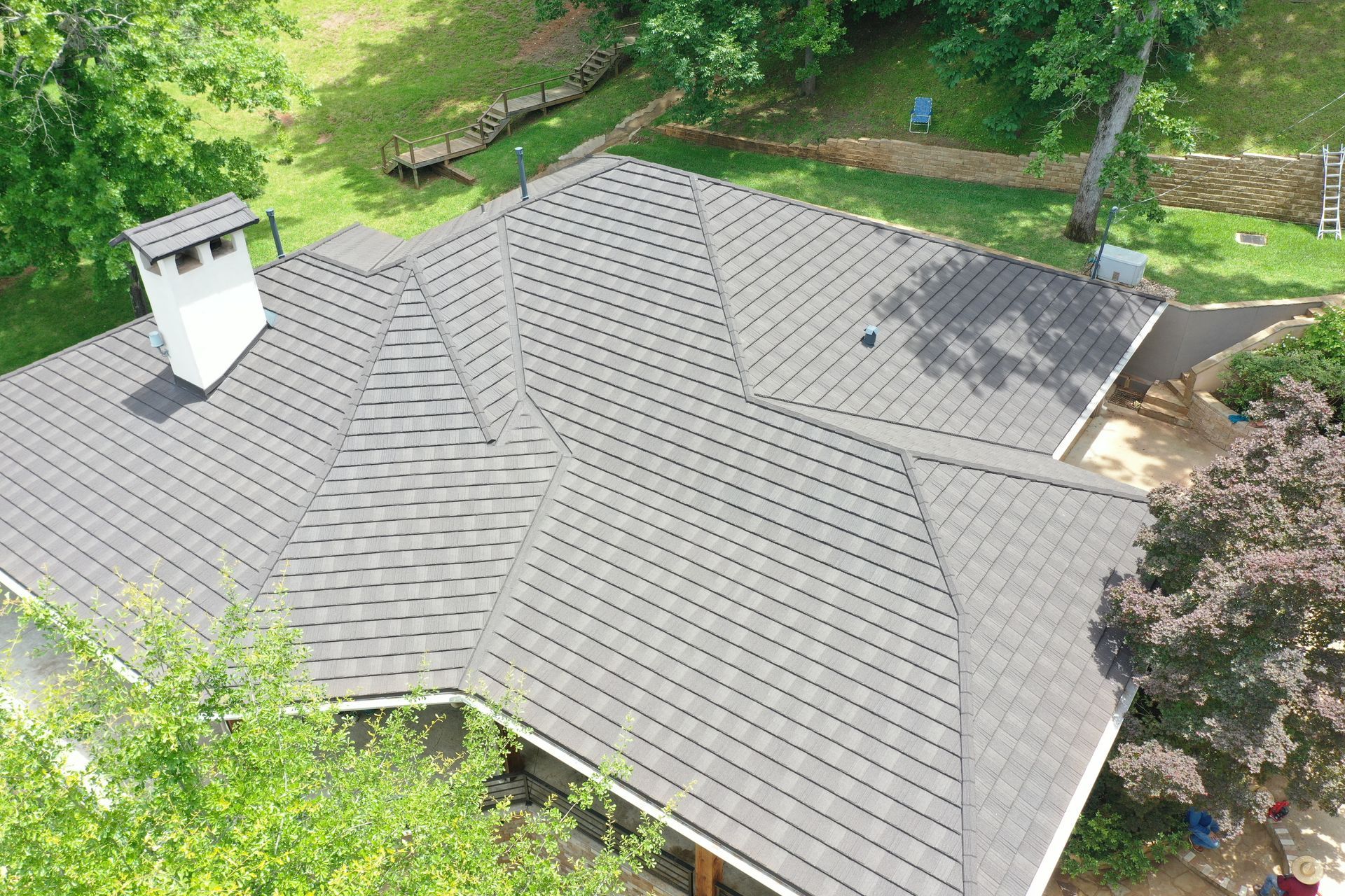 Aerial view of a gray shingled house roof with a white chimney, surrounded by green trees.