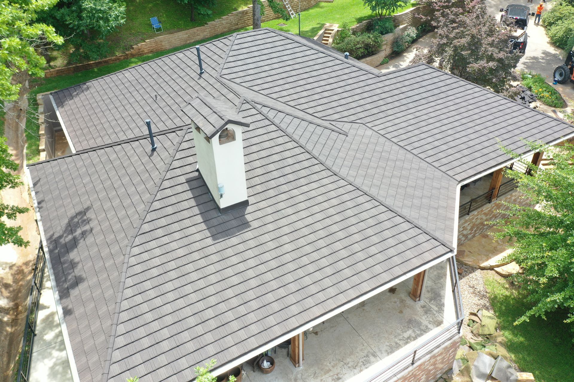 Aerial view of a gray-shingled house roof with a chimney, surrounded by trees and greenery