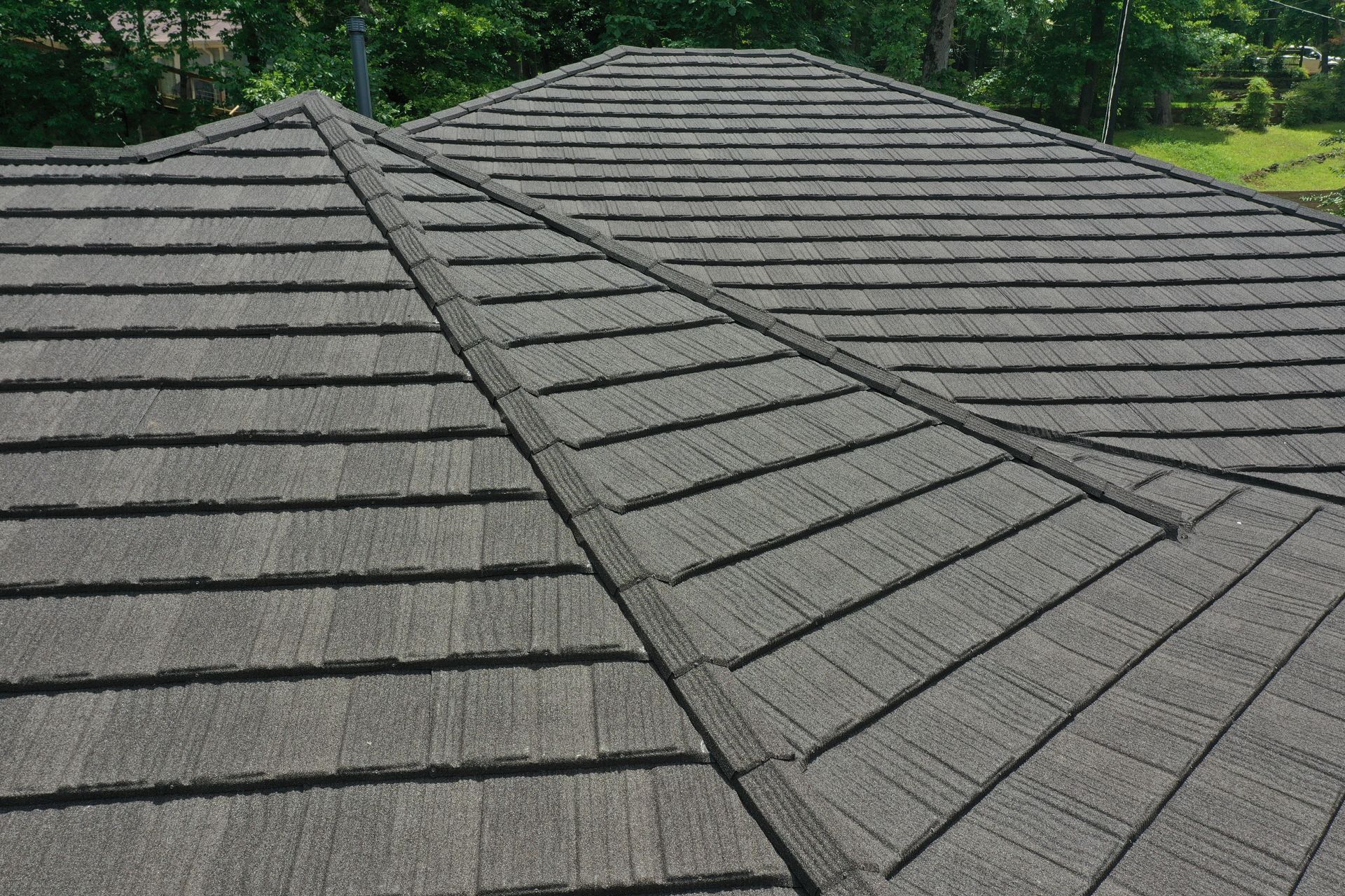 Gray shingled roof with intersecting ridges against green trees in the background
