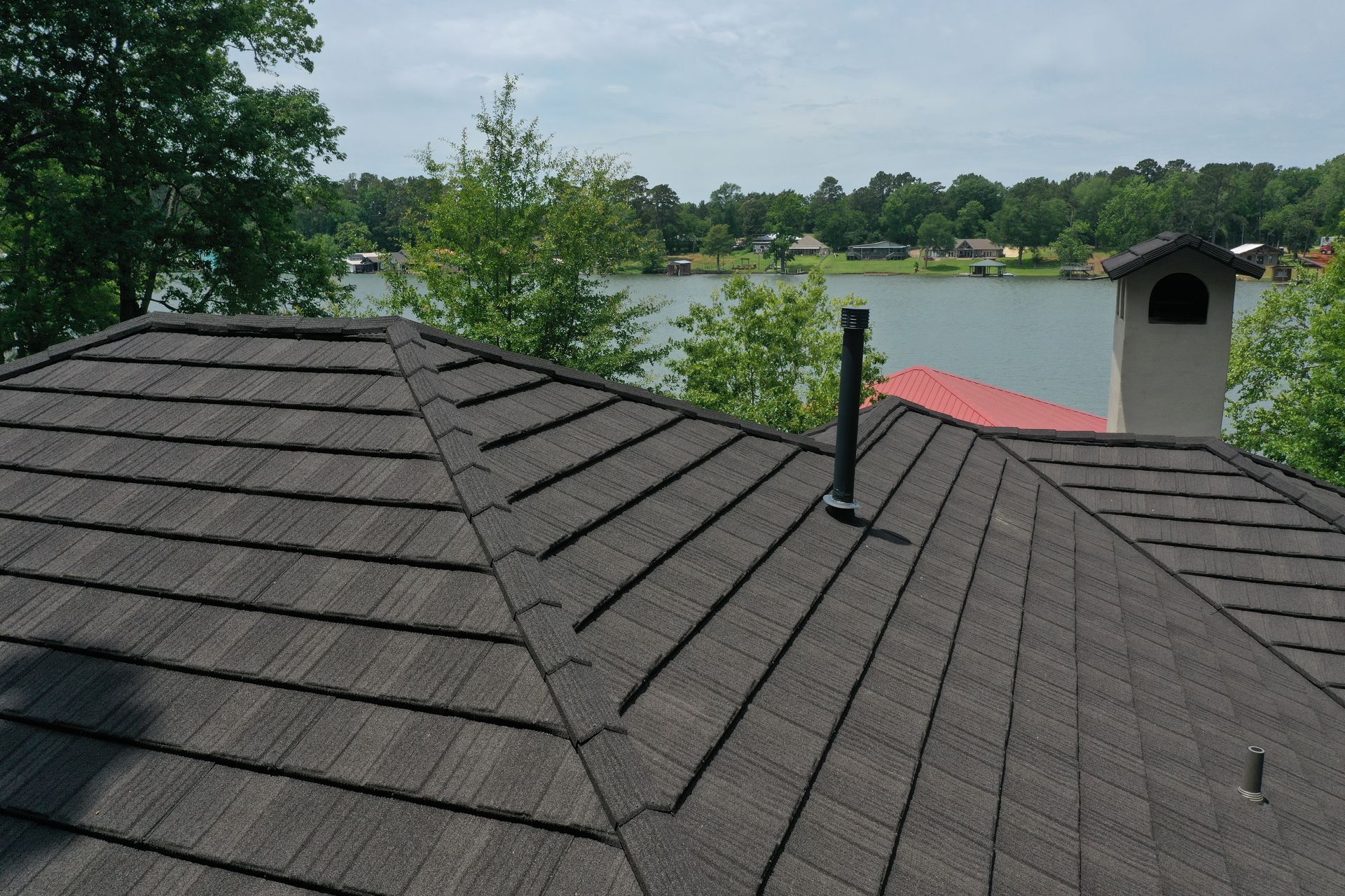 Dark shingled roof overlooking a lake, trees, and a chimney under a cloudy sky