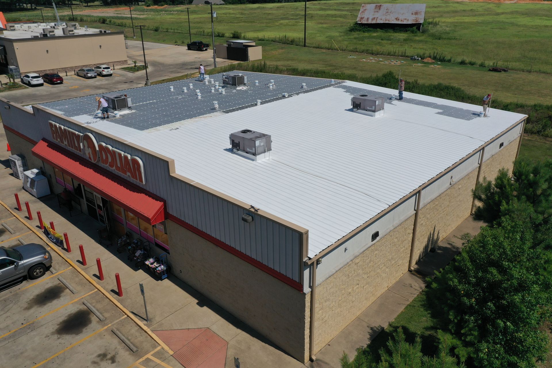 Aerial view of a beige commercial building with a white roof and red awnings beside a parking lot and green field