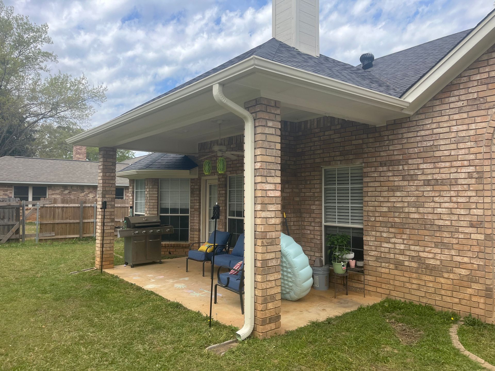 Covered patio beside a brick house with outdoor furniture, grill, and backyard lawn