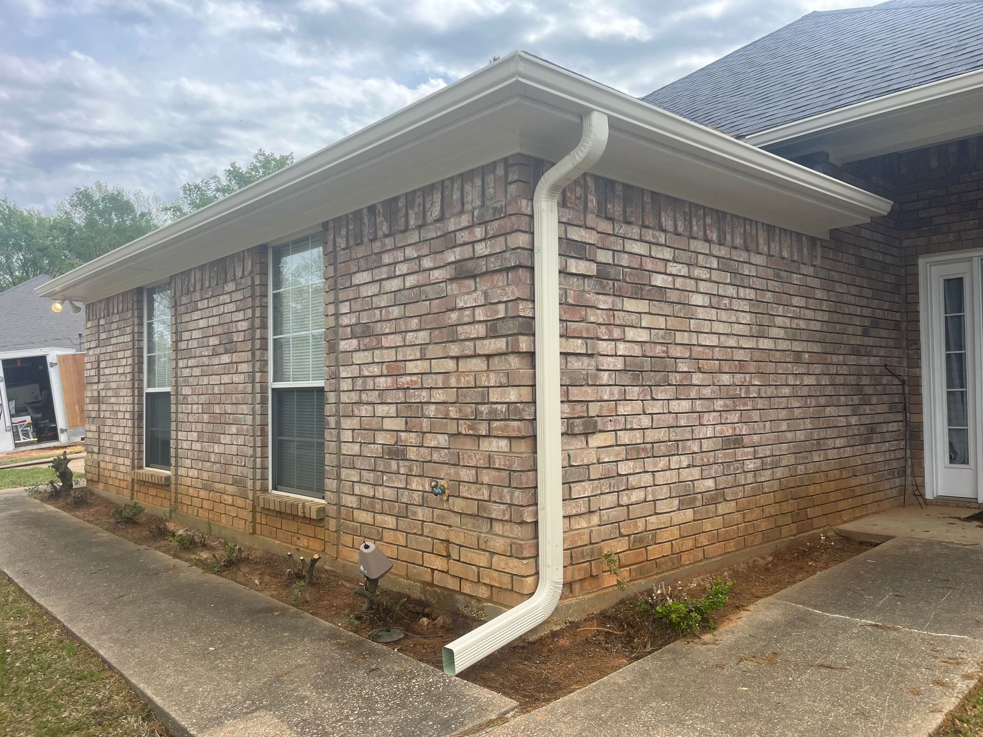 Brick house exterior with white gutter, windows, and concrete walkway under a cloudy sky