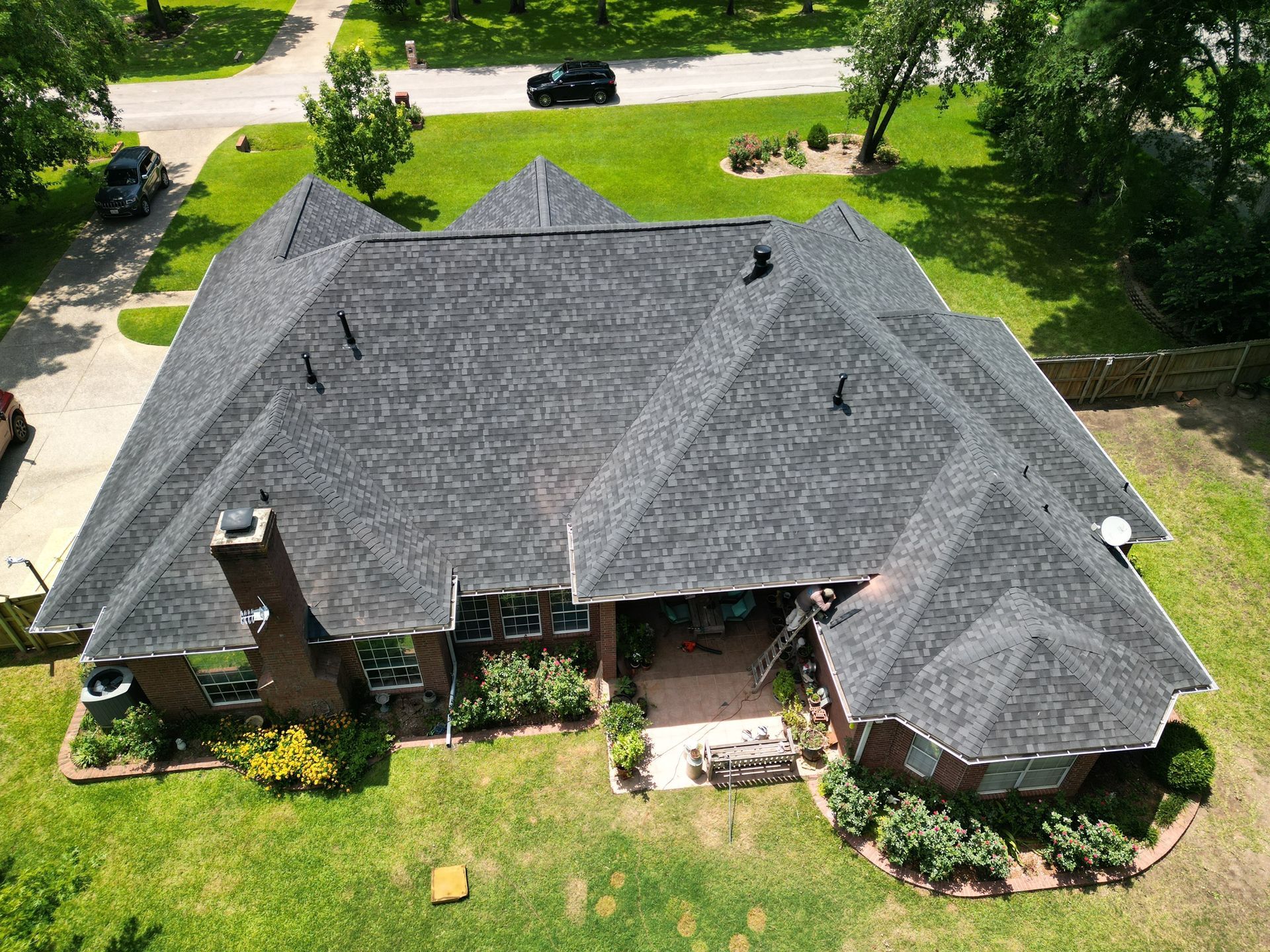 Aerial view of a brick house with a dark shingled roof and green lawn surrounded by trees.