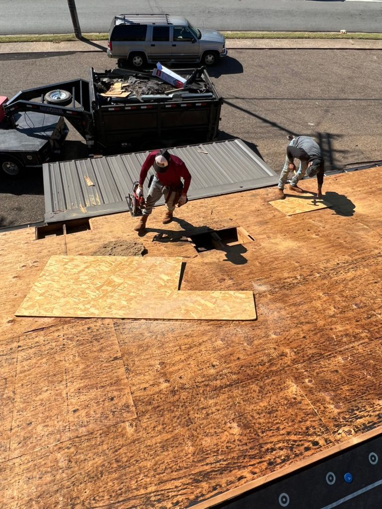 Two workers installing plywood sheets on a roof beside a truck with building materials.