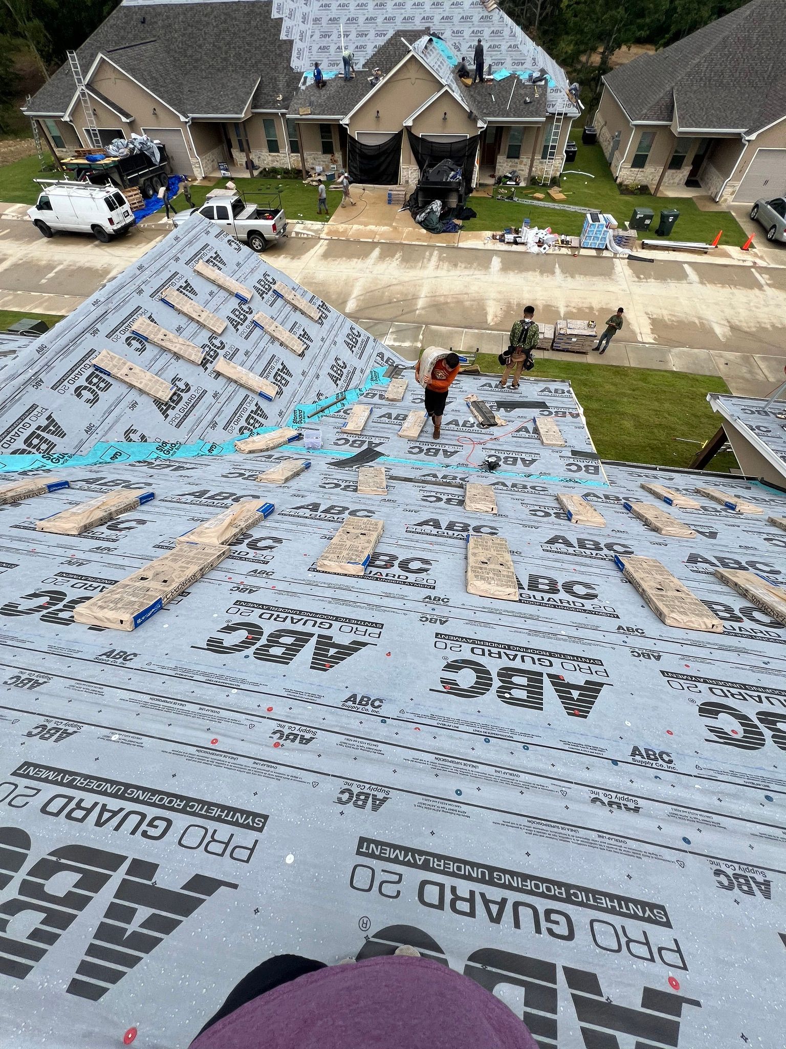 Worker on a steep roof covered in blue-gray underlayment, with houses and a front yard below.