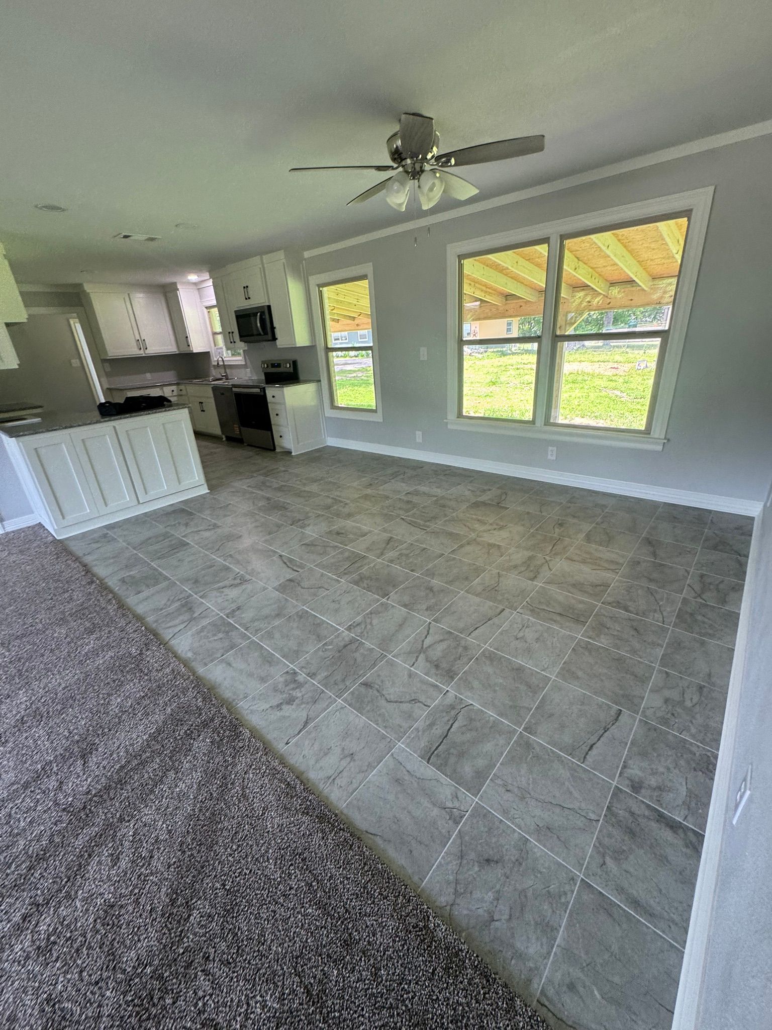 Empty living room with gray tile floor, ceiling fan, and large windows overlooking a yard.