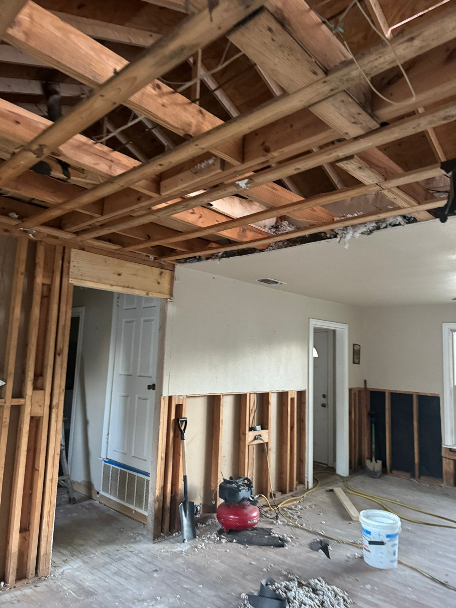 Partially demolished room with exposed ceiling joists, wall studs, debris, and tools during renovation
