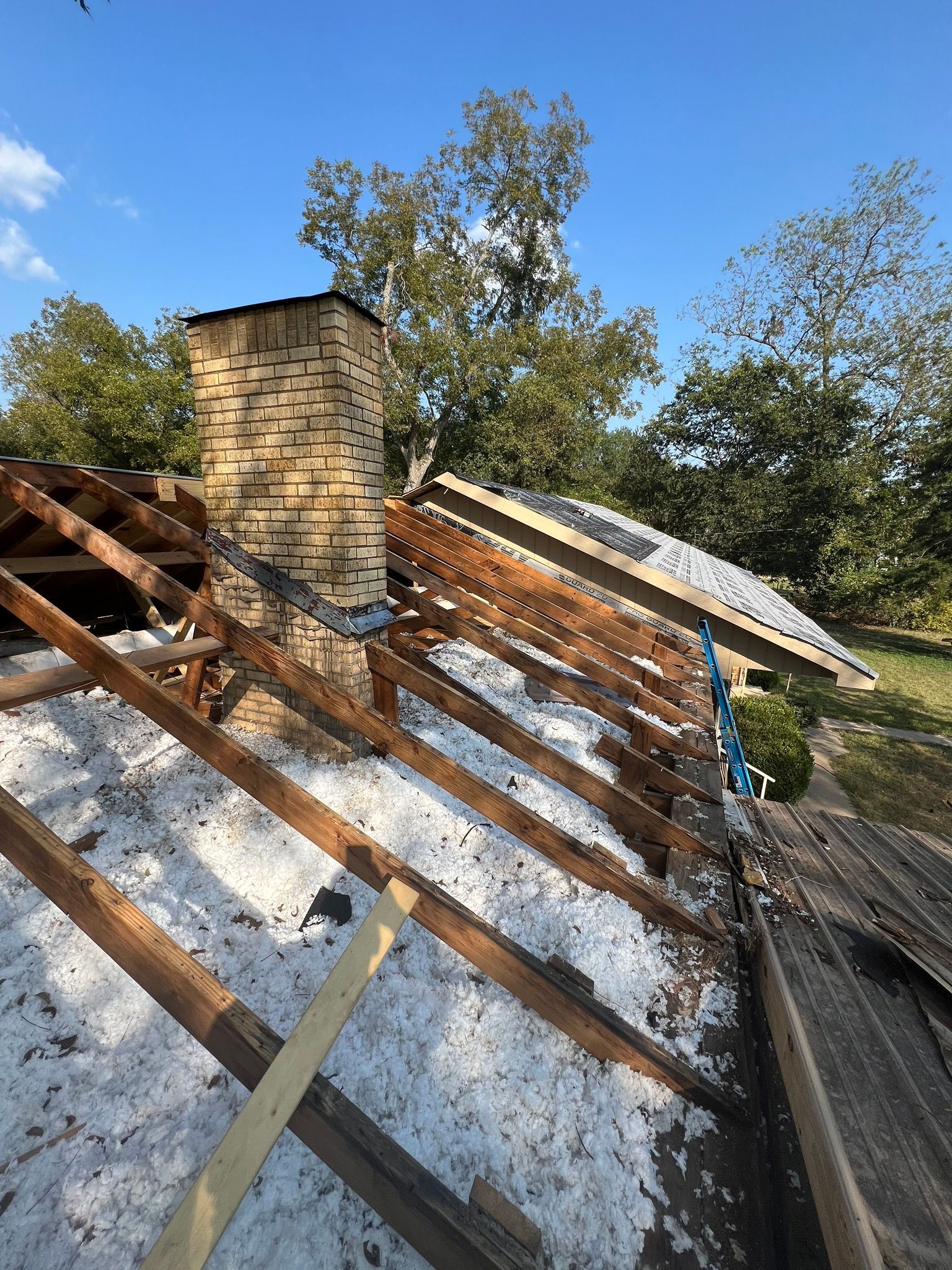 Damaged roof with exposed wooden rafters, a brick chimney, and patches of snow under a blue sky