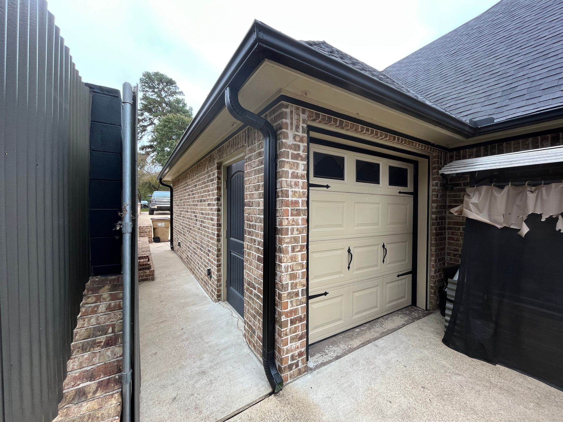 Narrow side driveway beside a brick garage with a beige door and dark roof, between a fence and house.