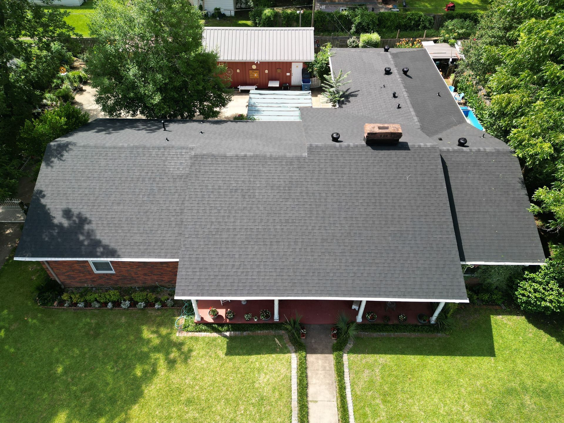 Aerial view of a house with a dark shingled roof, front porch, and green yard surrounded by trees.