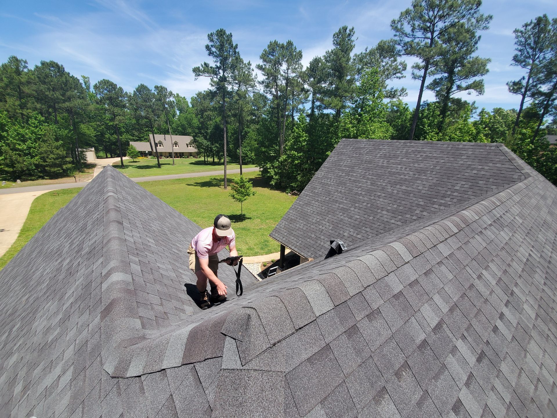 Person kneeling on a shingled roof, inspecting a roof valley in a suburban yard.