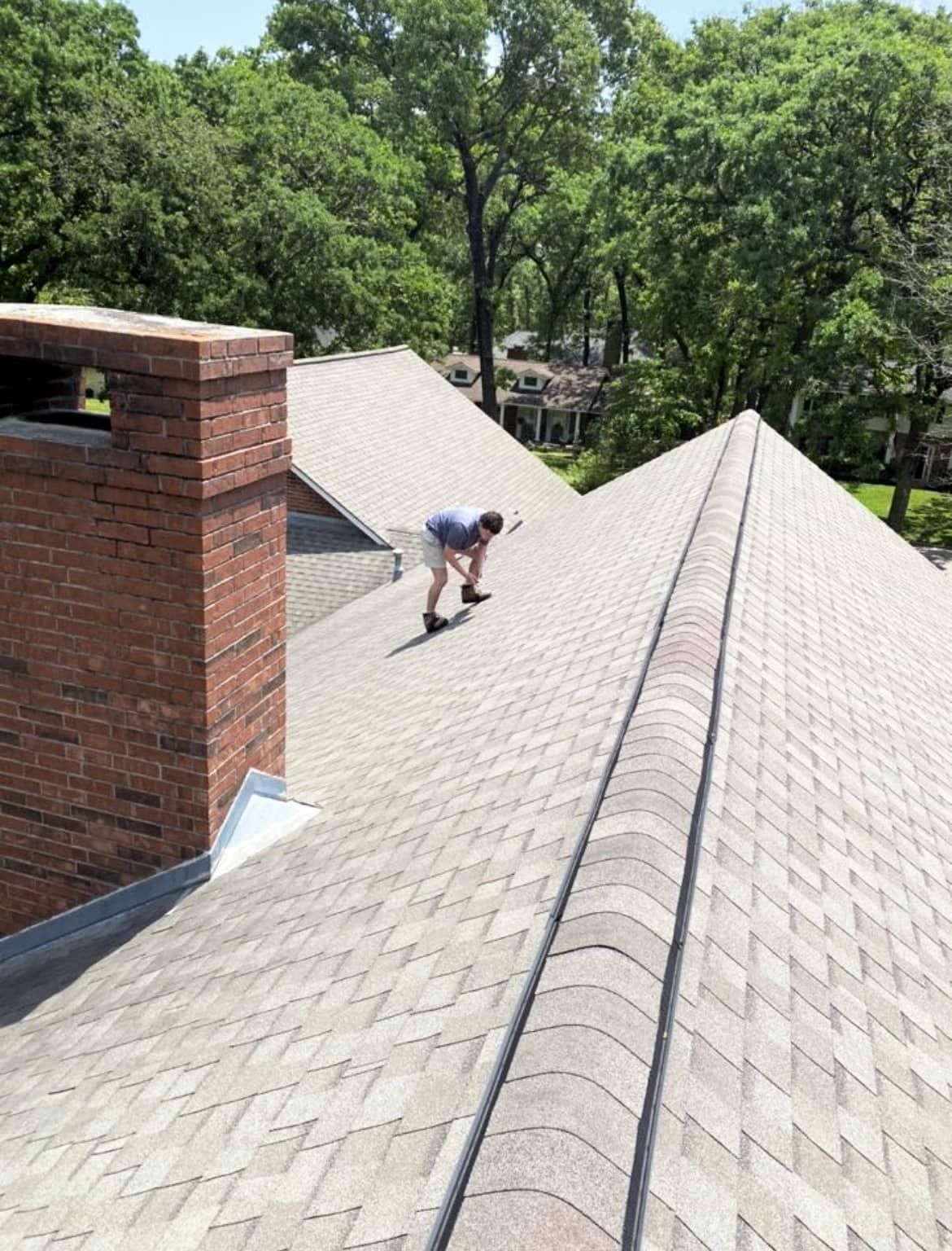 Worker on a shingled roof inspecting or repairing shingles near a brick chimney.