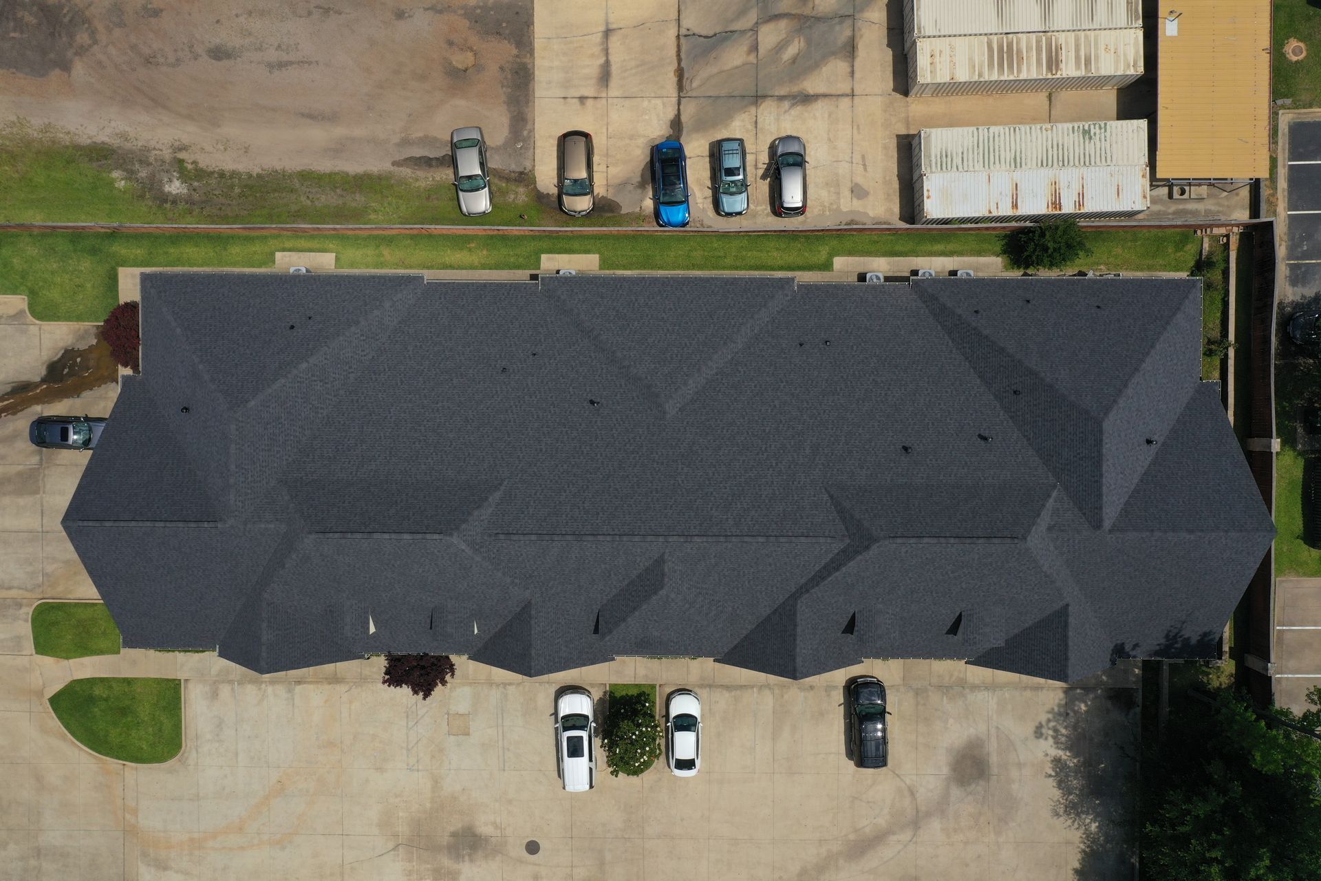 Aerial view of a dark gray roofed apartment building with parked cars and concrete lots below