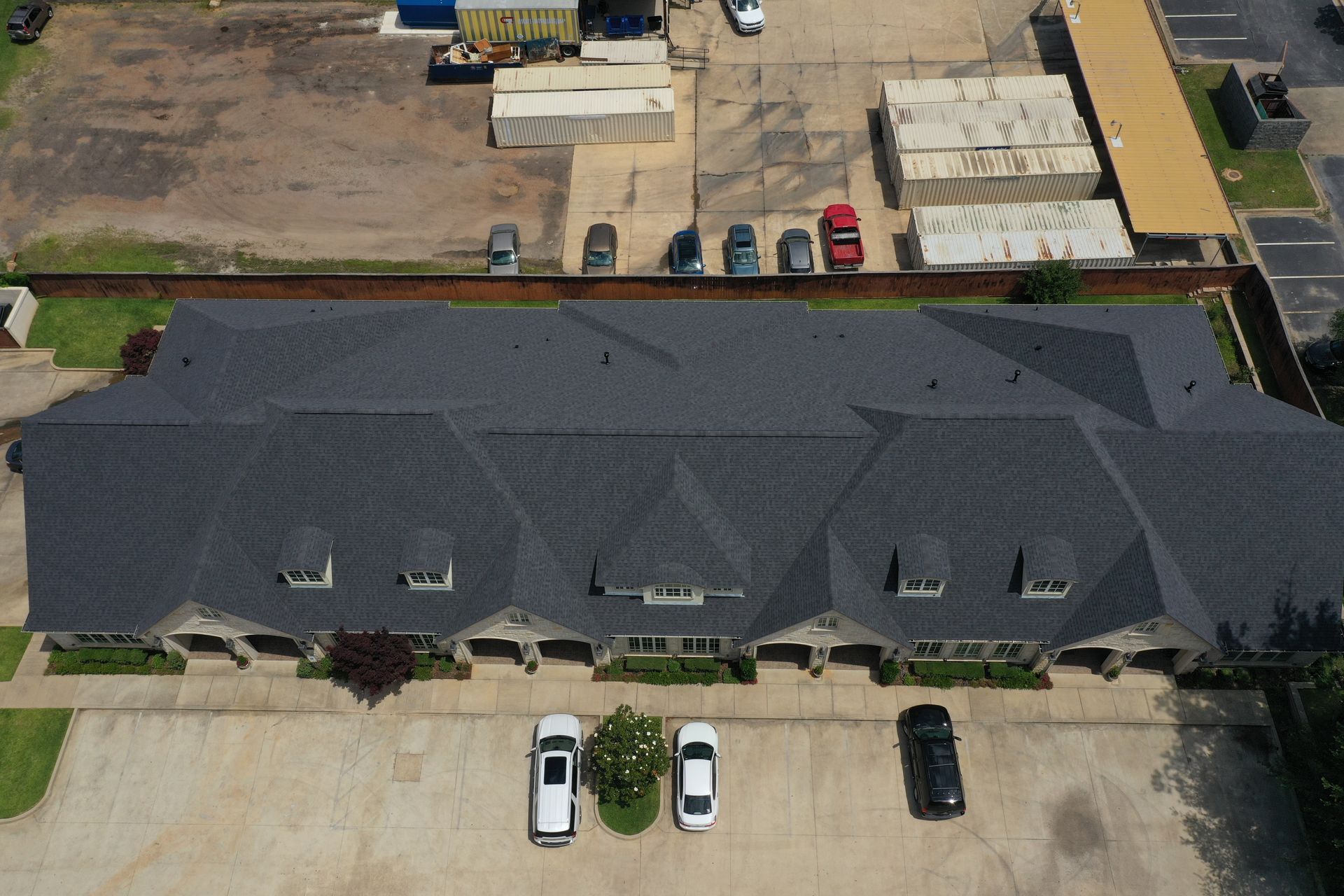 Aerial view of a long gray-roofed apartment building with parked cars in front and a lot behind.