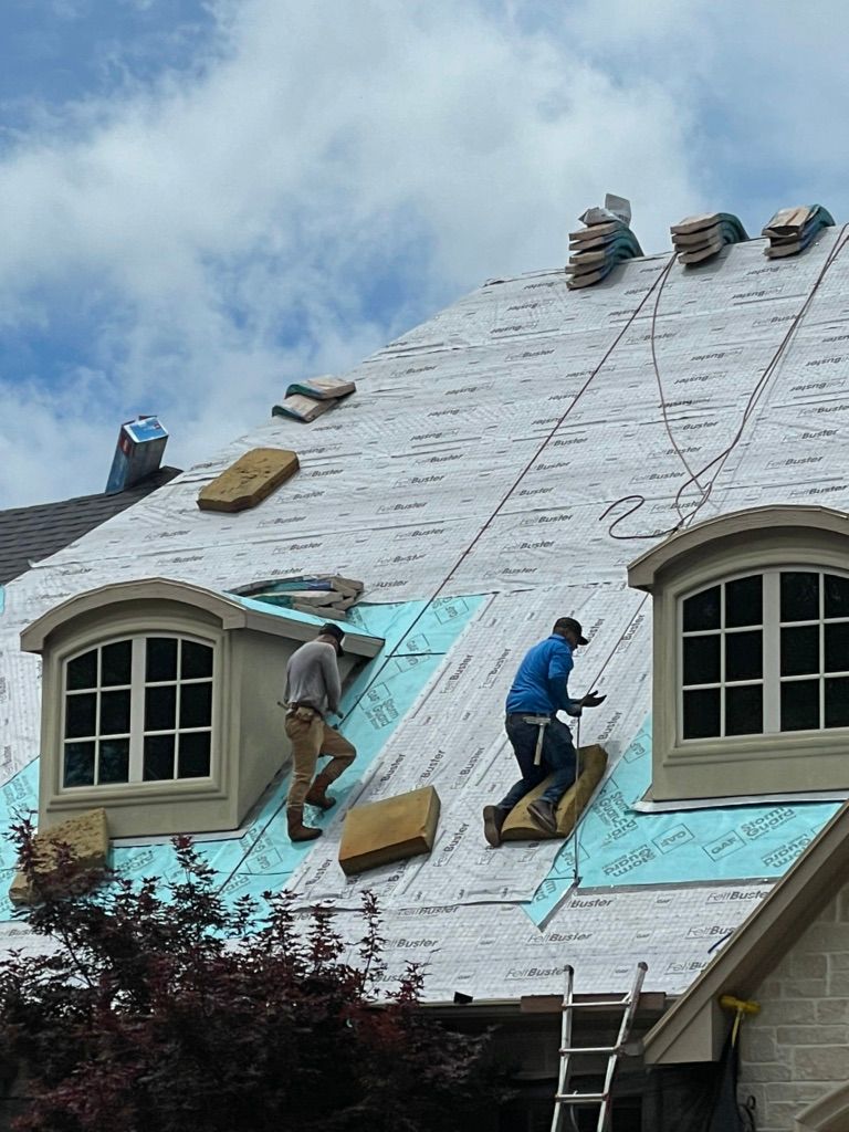 Roofers installing blue underlayment on a house roof, with ladders and windows below.