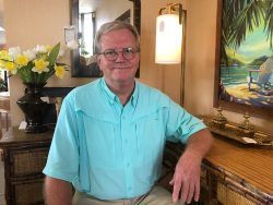 Man in a turquoise shirt seated indoors beside flowers and a framed beach painting.