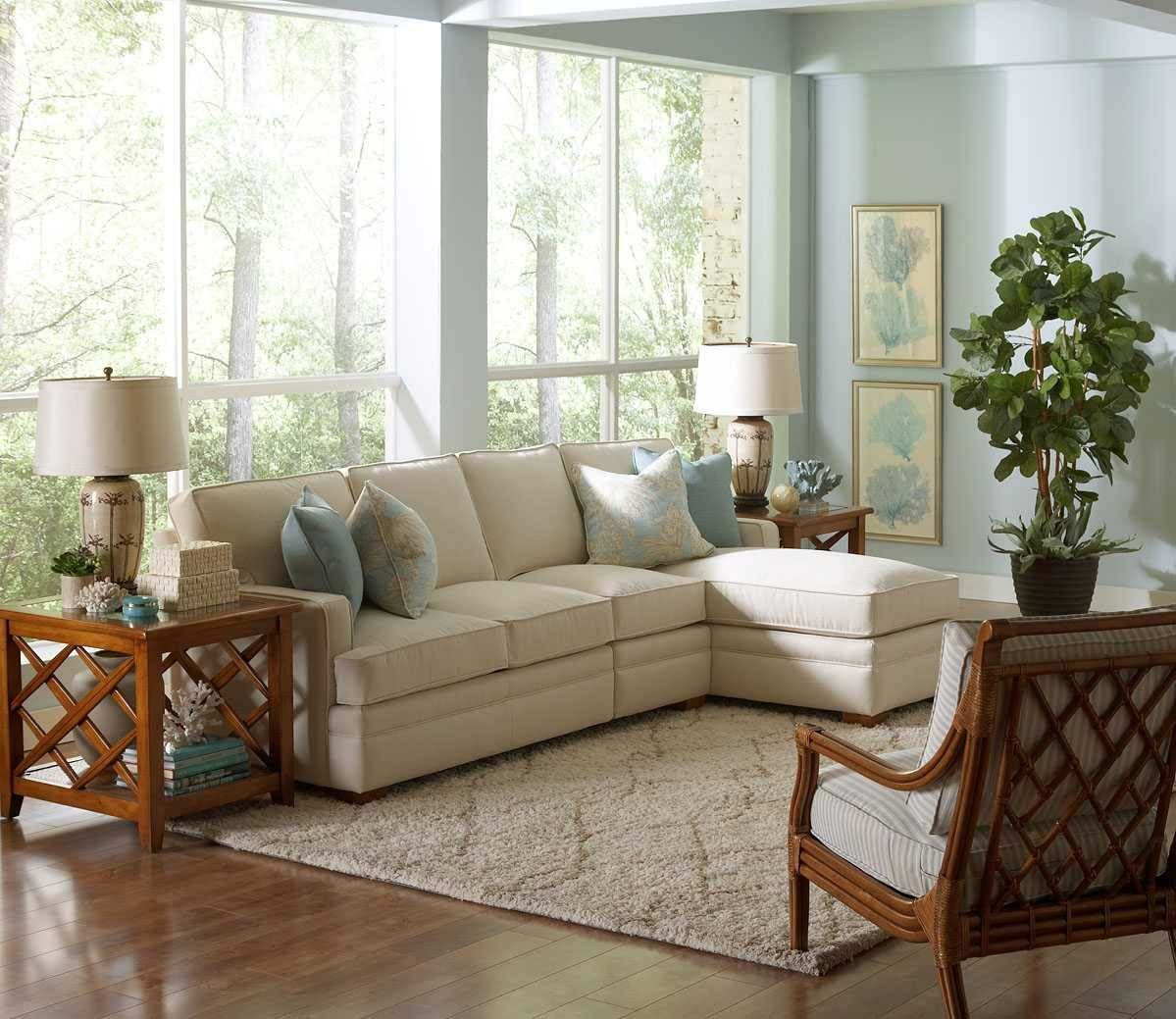Bright living room with beige sectional, wooden chairs, lamps, rug, and large windows with plants