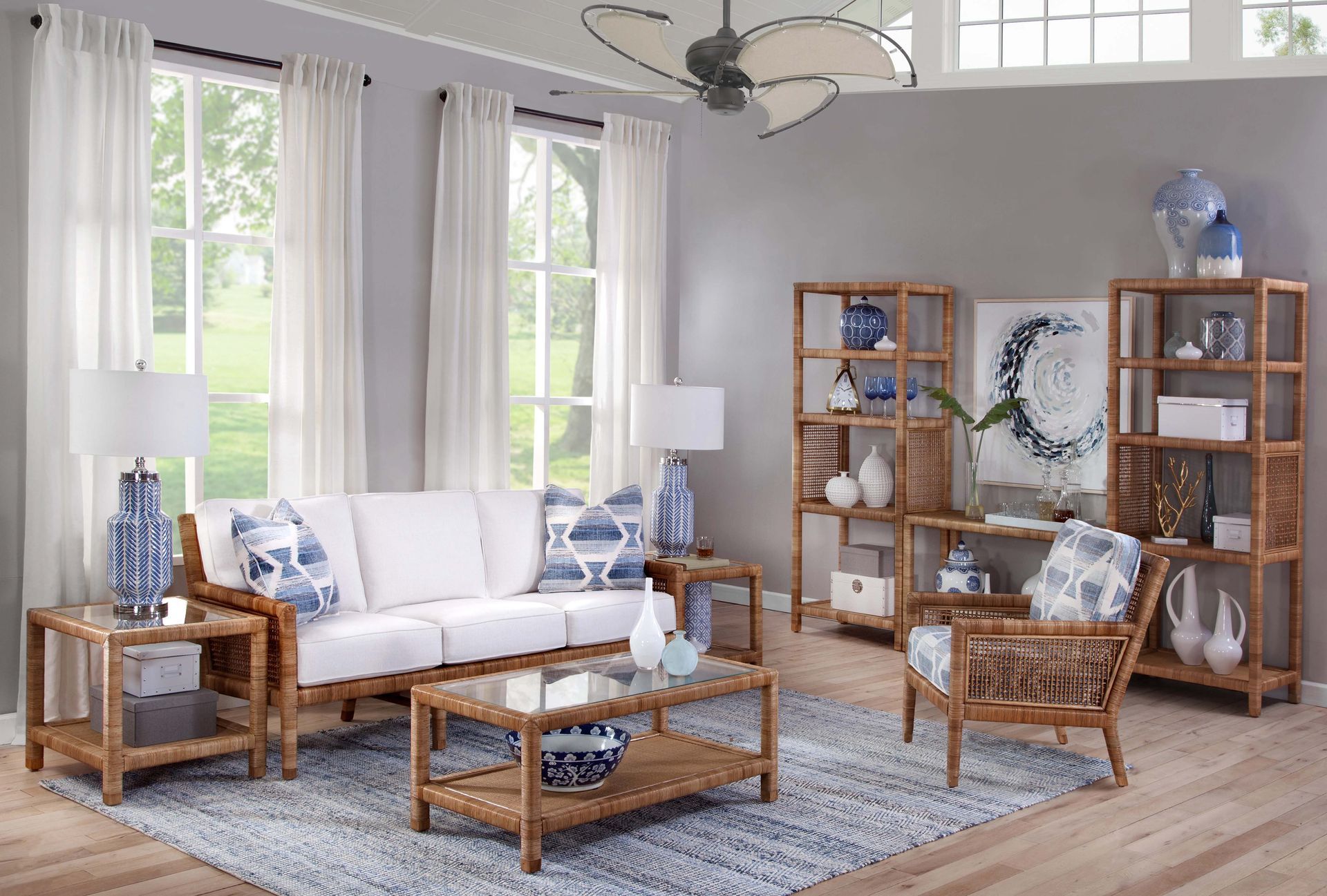 Bright coastal-style living room with white sofa, blue accents, rattan chairs, and large windows