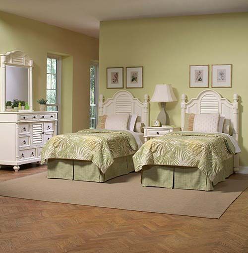 Twin white beds in a pale green bedroom with matching dresser, lamps, framed art, and tan rug