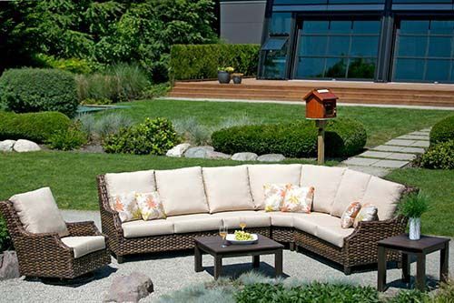 Outdoor patio seating with beige sectional sofa, wicker chair, and coffee tables in a landscaped backyard garden
