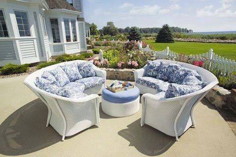 White outdoor wicker seating set with blue cushions around a round table on a sunny patio by a garden and fields
