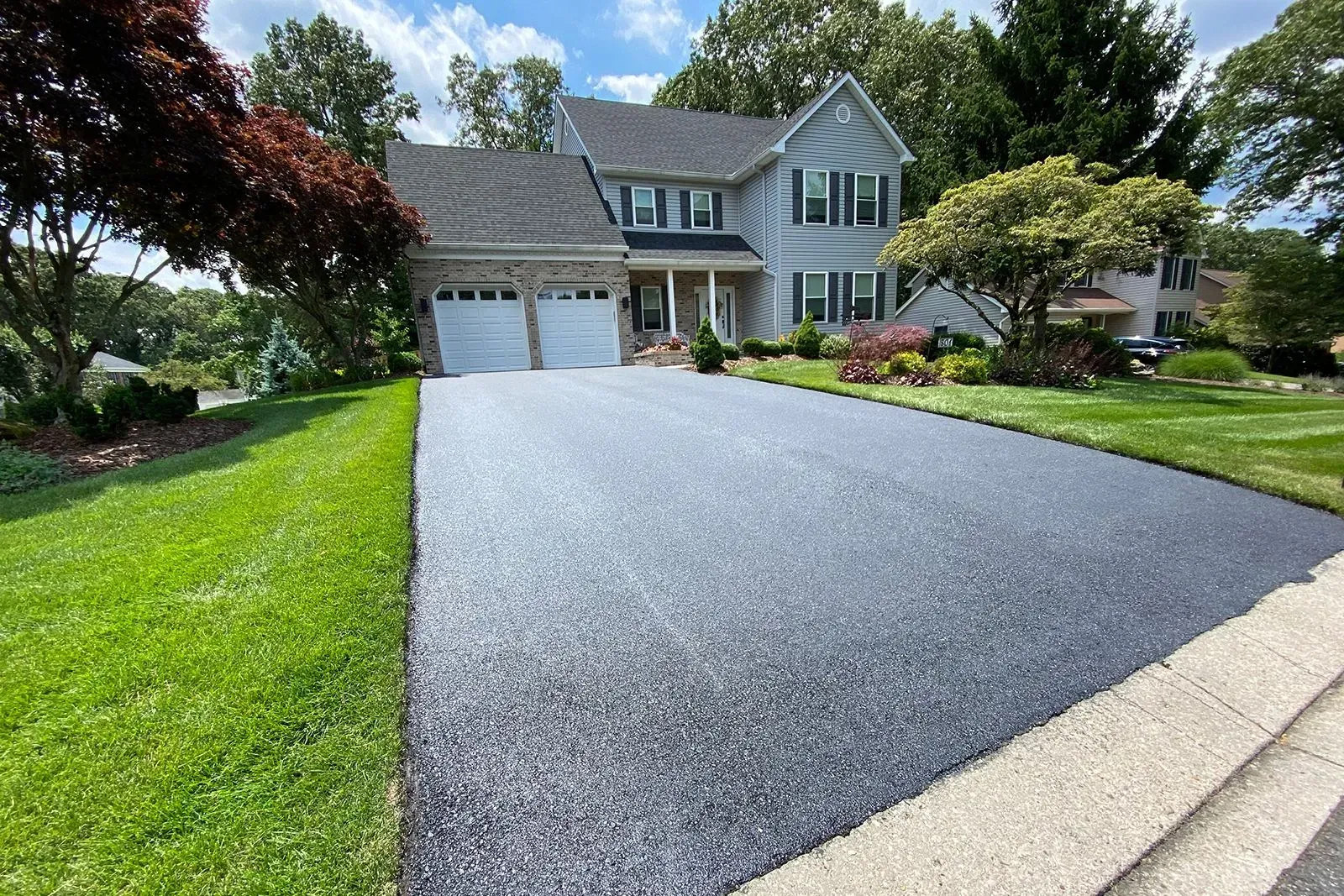 Black asphalt driveway leads to a two-story house with a green lawn. Sunny day.