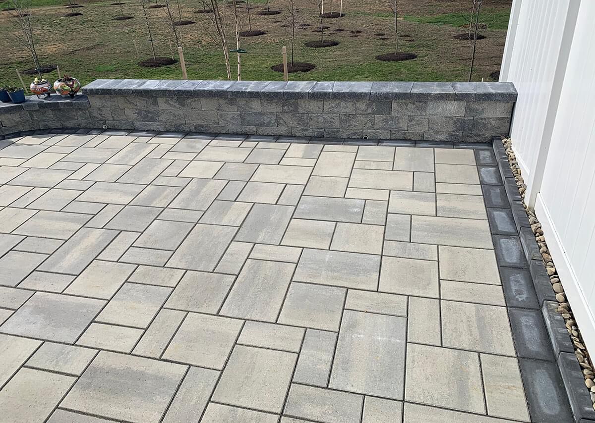 Gray paver patio with stone retaining wall and white siding.