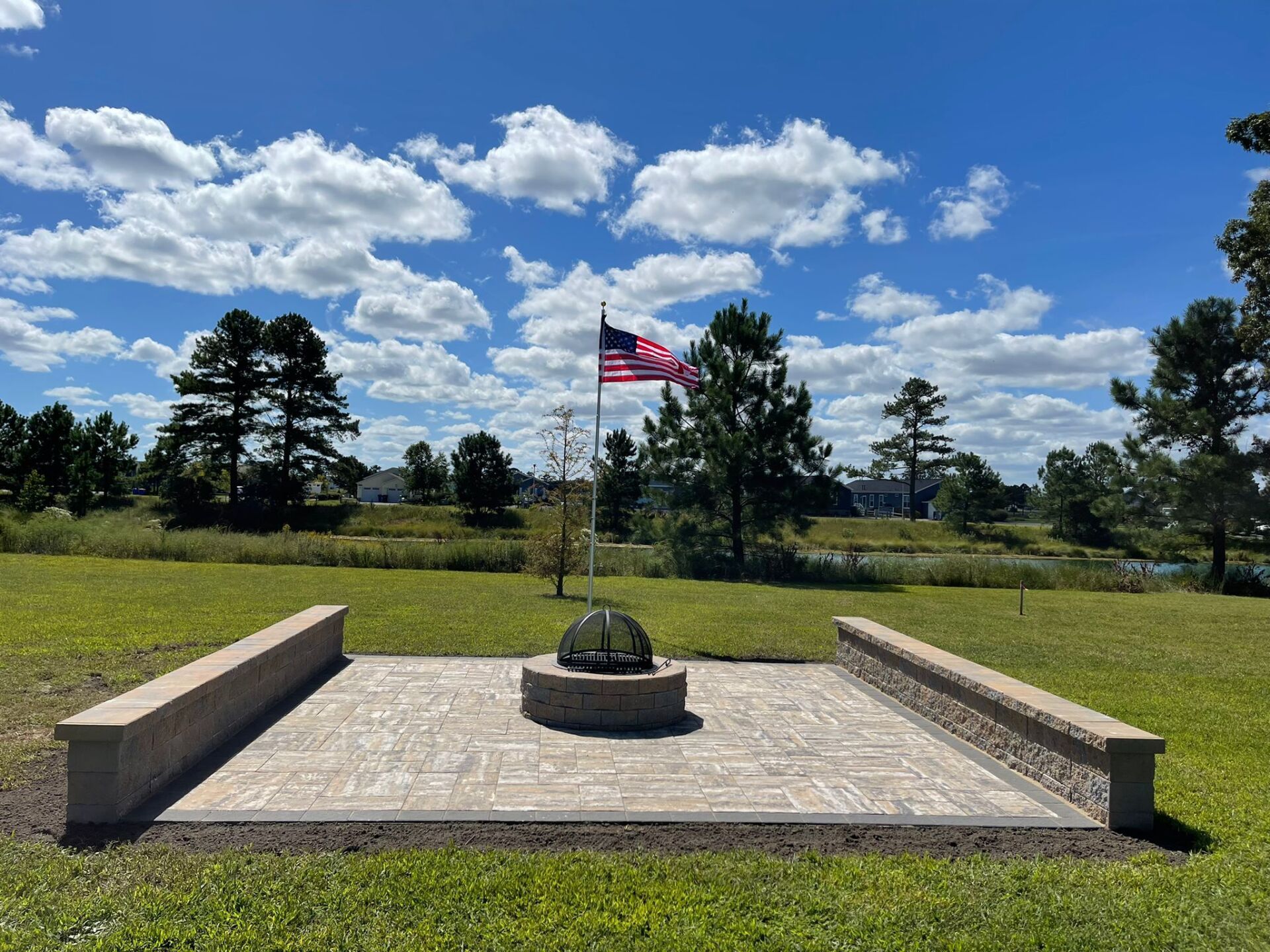 Patio with American flag, sphere sculpture, and low brick walls on a grassy landscape under a blue, cloudy sky.