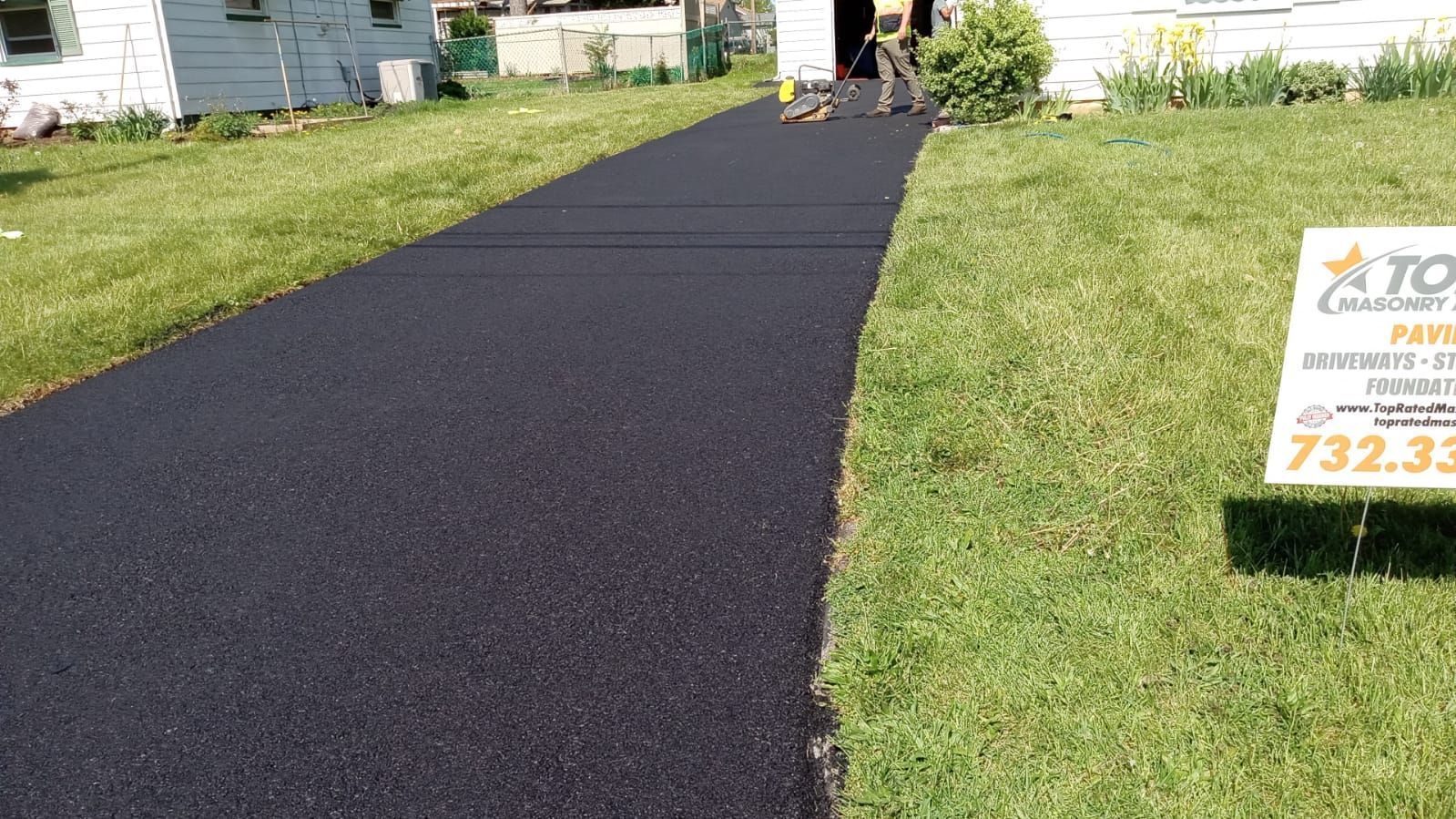 Newly paved black asphalt driveway next to green grass and a business sign.