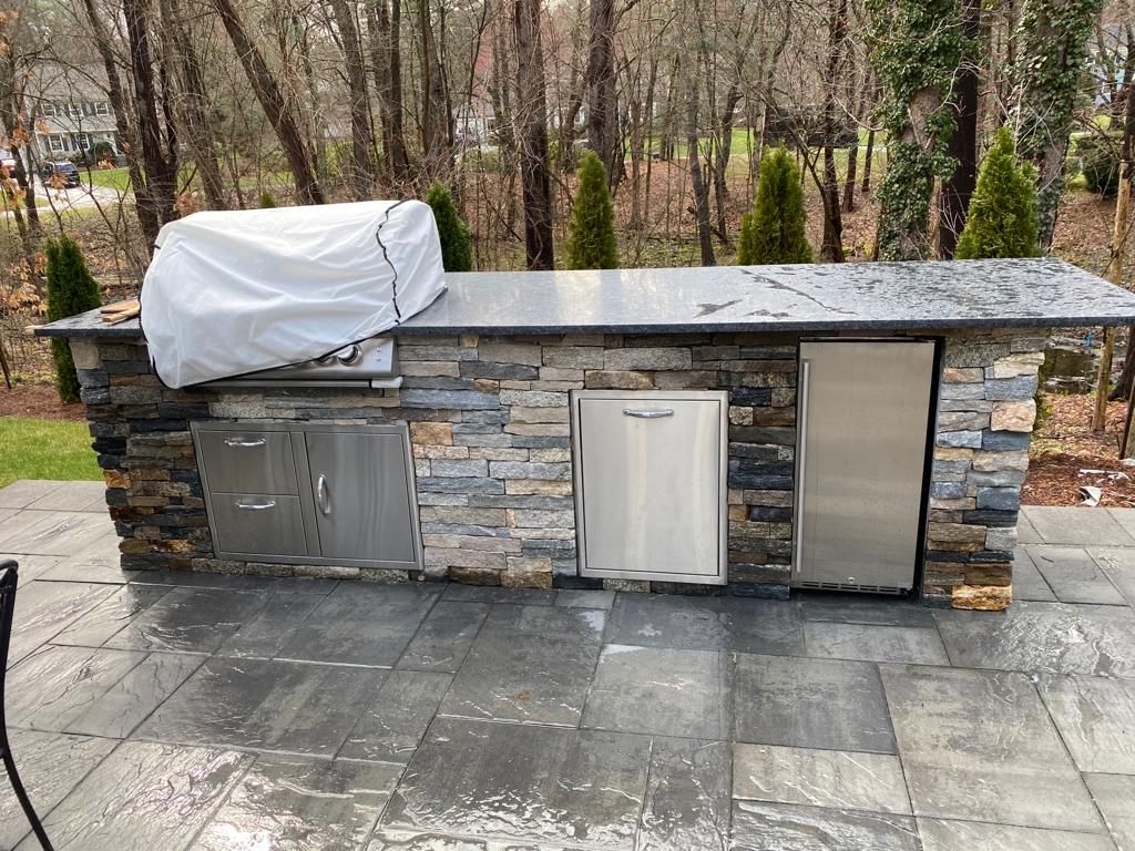 Outdoor kitchen with a covered grill, stone facade, and stainless steel appliances on a patio.