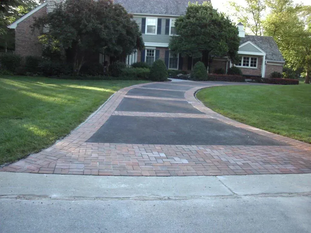 Driveway leading to a brick home. Driveway is a mix of brick and asphalt, surrounded by green grass.