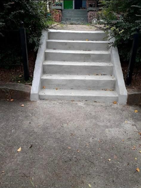 Concrete stairs leading up to a building entrance. The stairs are flanked by dark posts and overgrown bushes.