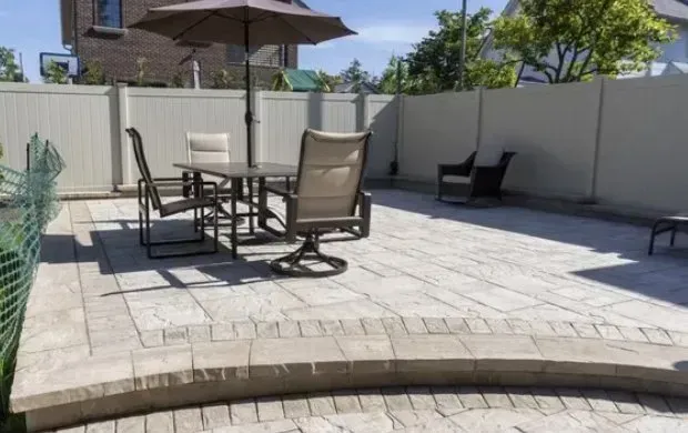 Patio with outdoor furniture, tan pavers, and a white fence on a sunny day.