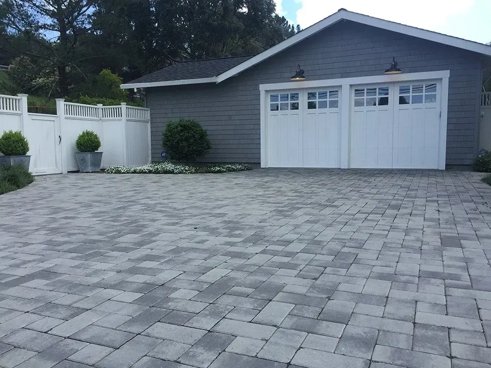 Brick driveway leading to a white garage with two doors, gray siding and fencing, green plants and blue sky.