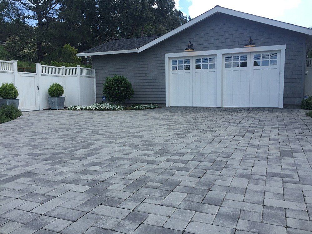 Gray brick driveway leading to a two-car garage with white doors, a white fence, and manicured landscaping.