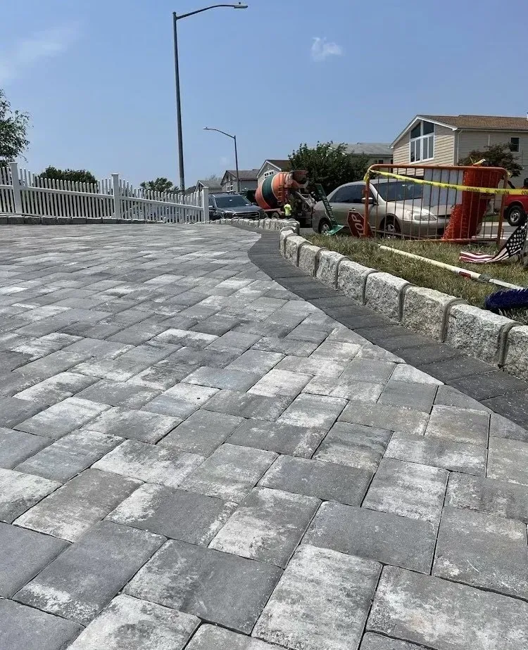 Paved driveway with gray pavers, bordered by a concrete curb. Construction equipment and houses in the background under a blue sky.