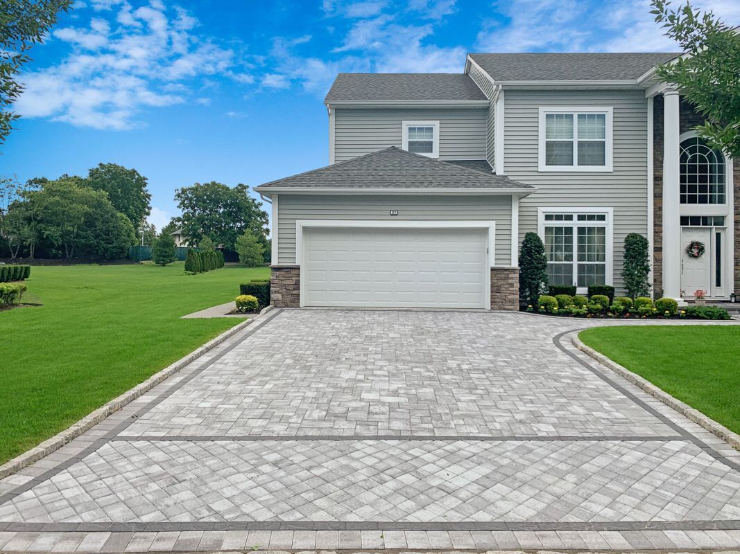 Two-story gray house with a brick facade and a paved driveway, framed by a lush green lawn and cloudy sky.