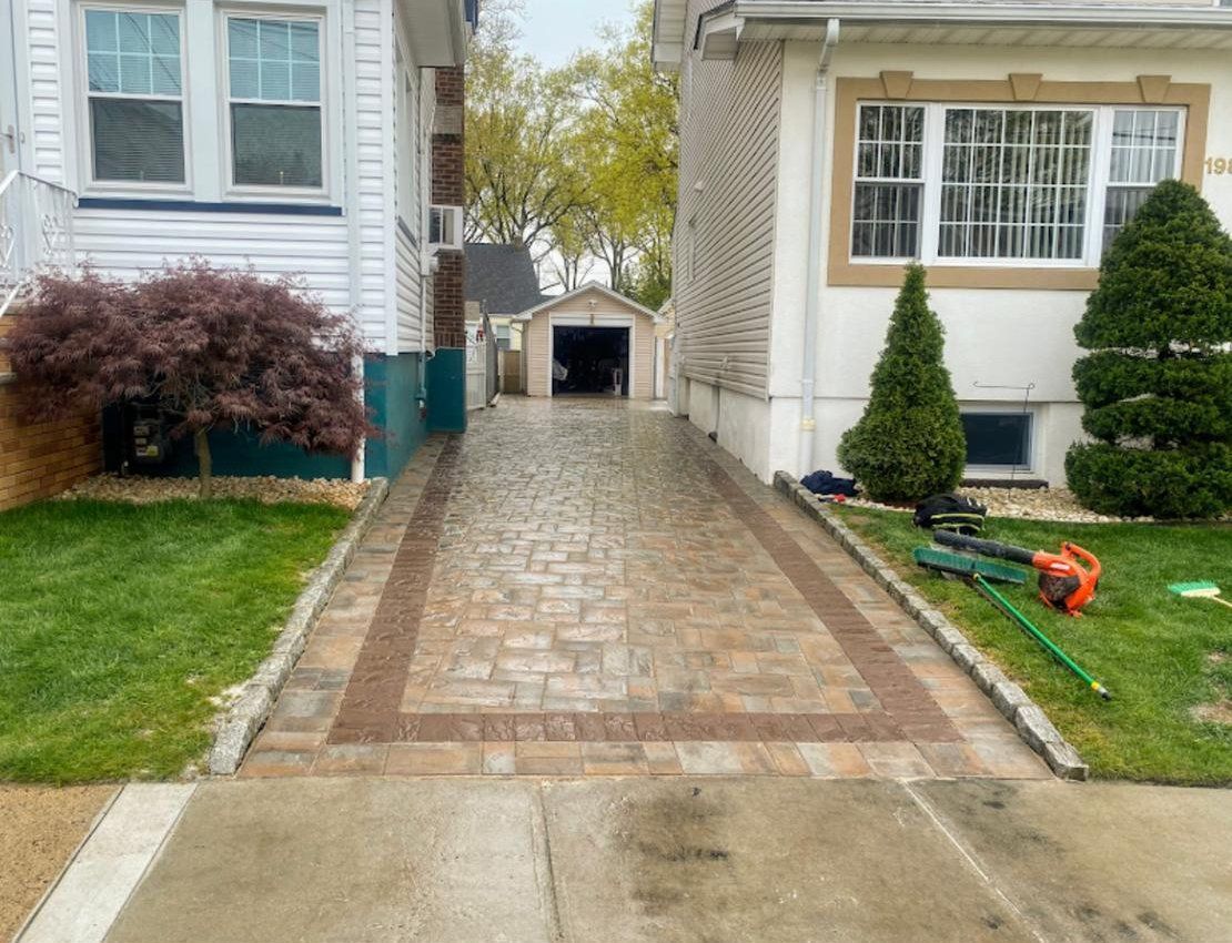 Brick driveway between two houses, with a garage visible in the distance. Lawn and shrubs on either side.