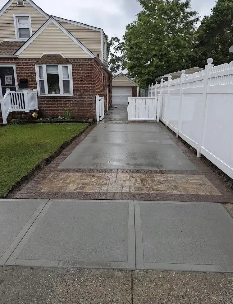 Driveway leading to a garage, bordered by brick and concrete. White fence on the right. House on the left.